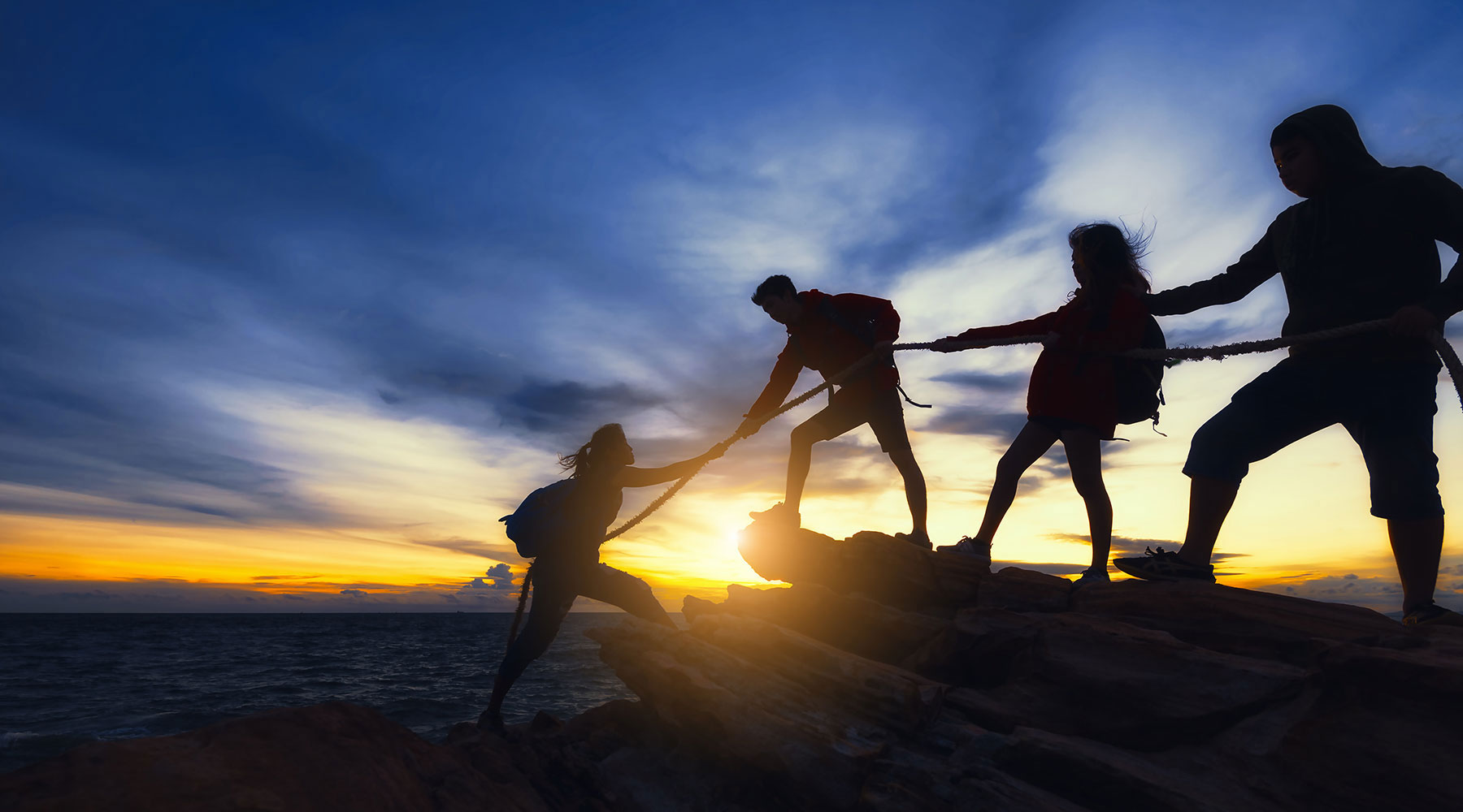 Silhouette of people holding hands on top of a rock at sunset, united by a rope.