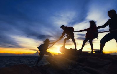 Silhouette of people holding hands on top of a rock at sunset, united by a rope.